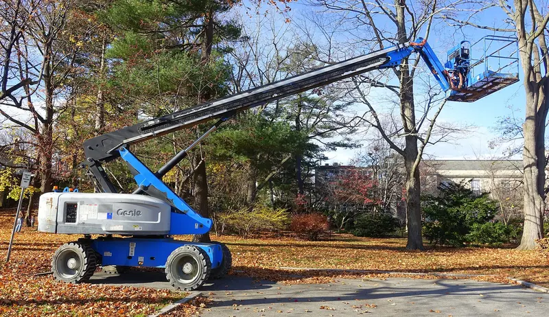 A blue Genie telescopic boom lift with its basket raised among trees beside a building.